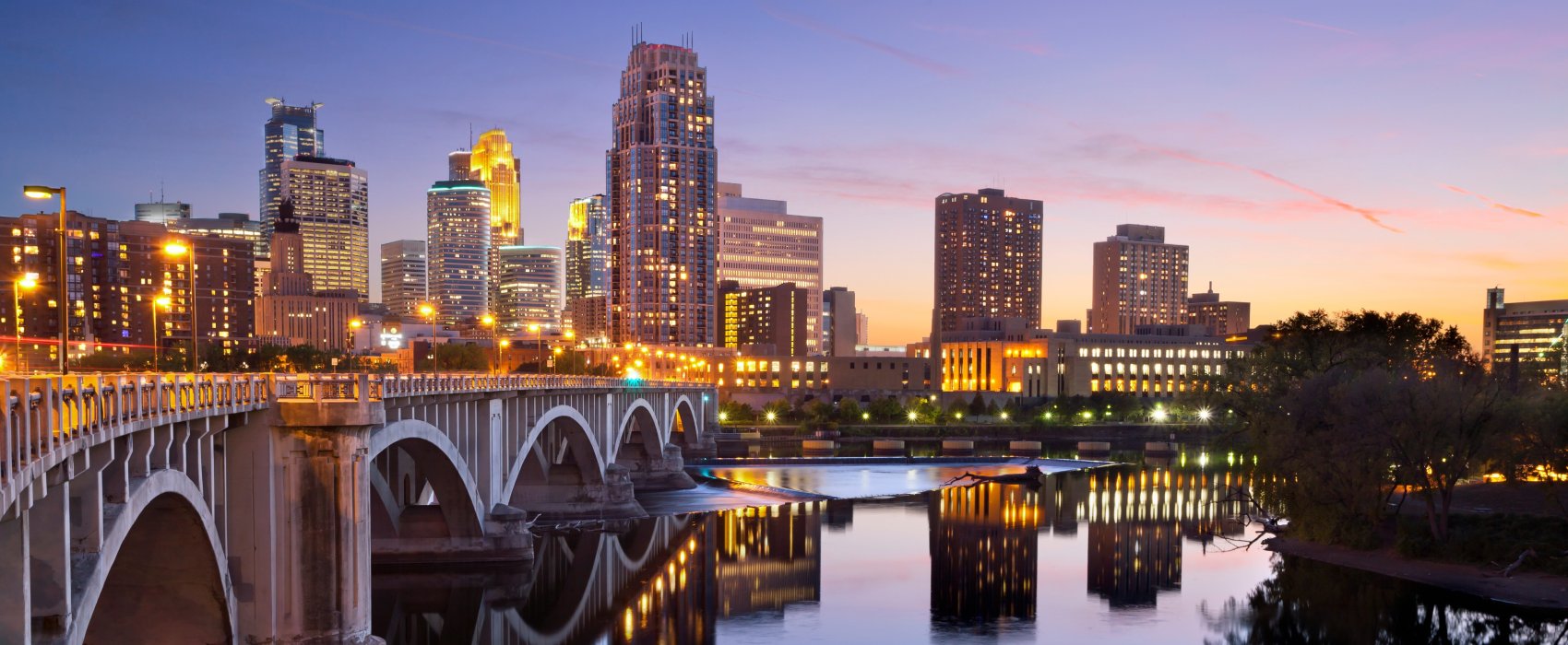 Minneapolis skyline in the background, river in the foreground. A purple sunset behind.