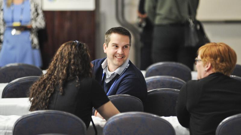 a smiling man turns aroudn in his chair to greet 2 other conference attendees at MES 2019